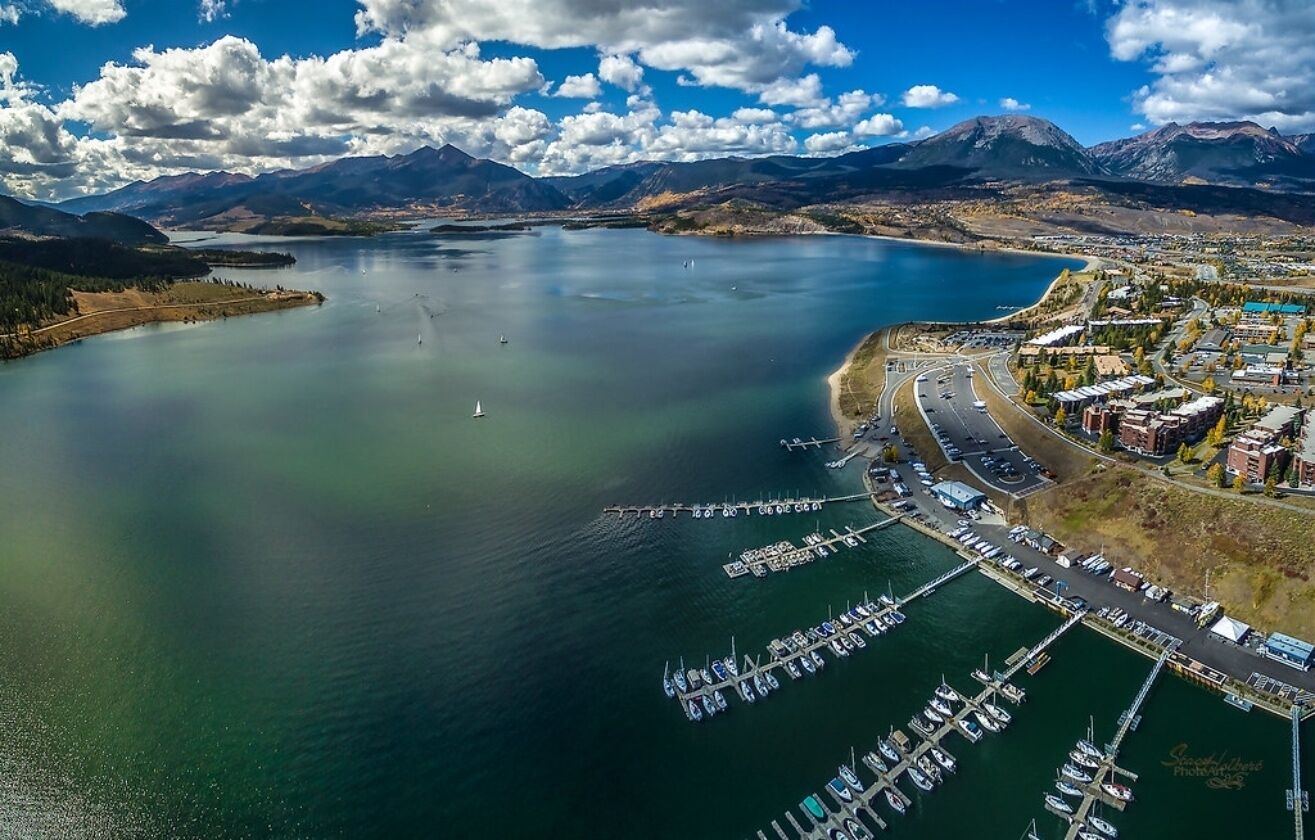 An aerial view of a coastal town with a marina, boats, and mountains in the background.