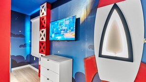 A room with a television mounted on the wall, a white dresser, and a red and blue wall design. - Luxury 8BR Themed Villa With Private Pool (Clermont)