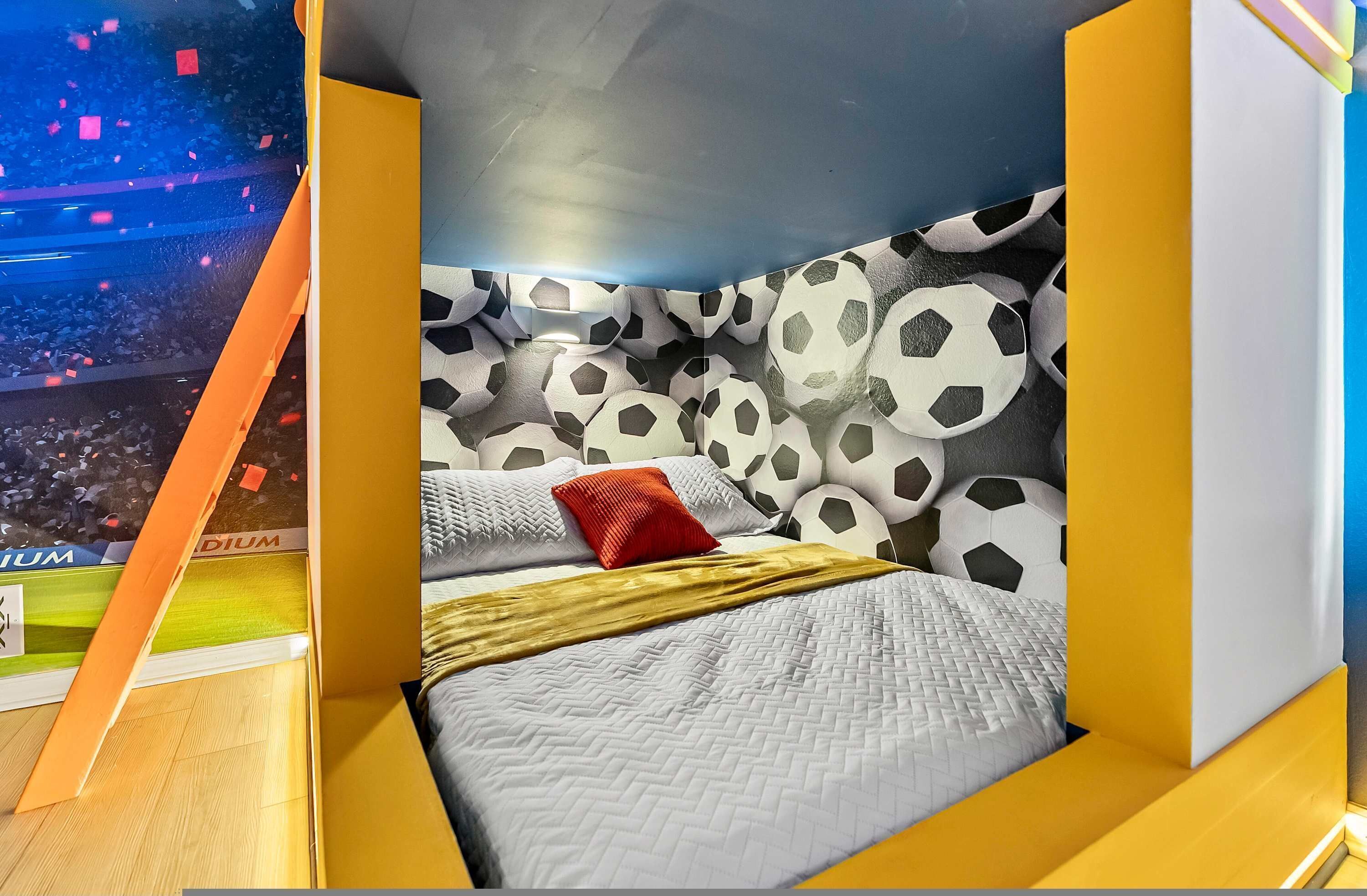Une chambre avec un lit dont la tête de lit est ornée d’un motif représentant un ballon de football, un oreiller rouge et une couverture jaune.