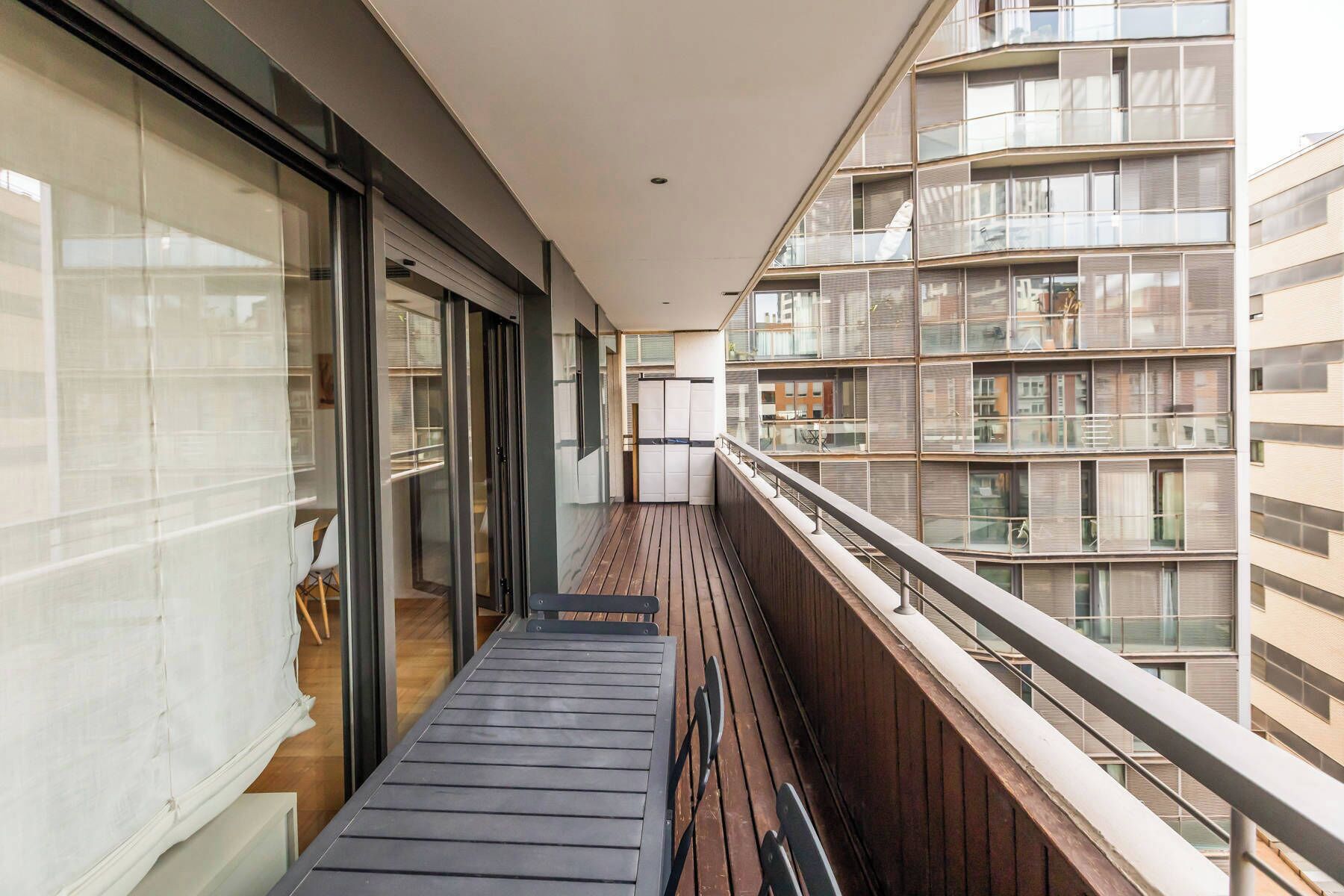 Een smal balkon met houten vloer, glazen deuren en uitzicht op omliggende gebouwen.