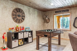 A room with a wooden foosball table, a large wall clock, a mounted deer head, and a window with a view of trees. - Aspen Pond Retreat | 7 Mi to YNP | Private Pond (West Yellowstone)