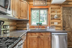 A rustic kitchen with wooden cabinets, a stainless steel sink, and a window with a view of greenery. - Aspen Pond Retreat | 7 Mi to YNP | Private Pond (West Yellowstone)