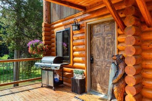 Une cabane en bois avec un barbecue sur la terrasse, une porte ornée d’une tête d’animal décorative et un panier à fleurs suspendu.