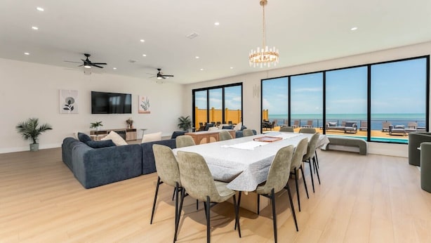 A spacious living area with a dining table, sofas, and a large window overlooking the sea. - NEW Luxury 5,500 SF Waterfront Home w/ Pool, Sleeps 28 (Port Isabel)