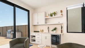 A modern kitchen with white cabinets, a built-in oven, and a sink. There are two chairs and a small table in the living area. - NEW Luxury 5,500 SF Waterfront Home w/ Pool, Sleeps 28 (Port Isabel)