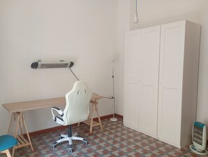 Huoneessa on työpöytä, valkoinen toimistotuoli, lattiavalaisin, vaatekaappi ja lattialla lämmitin.