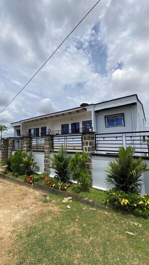 A modern two-story house with a stone wall and metal railing, surrounded by greenery and flowers. - Furnished Studio - Condo - WILMA HOUSE Furnished Apartments - YASSA - DOUALA (Douala)