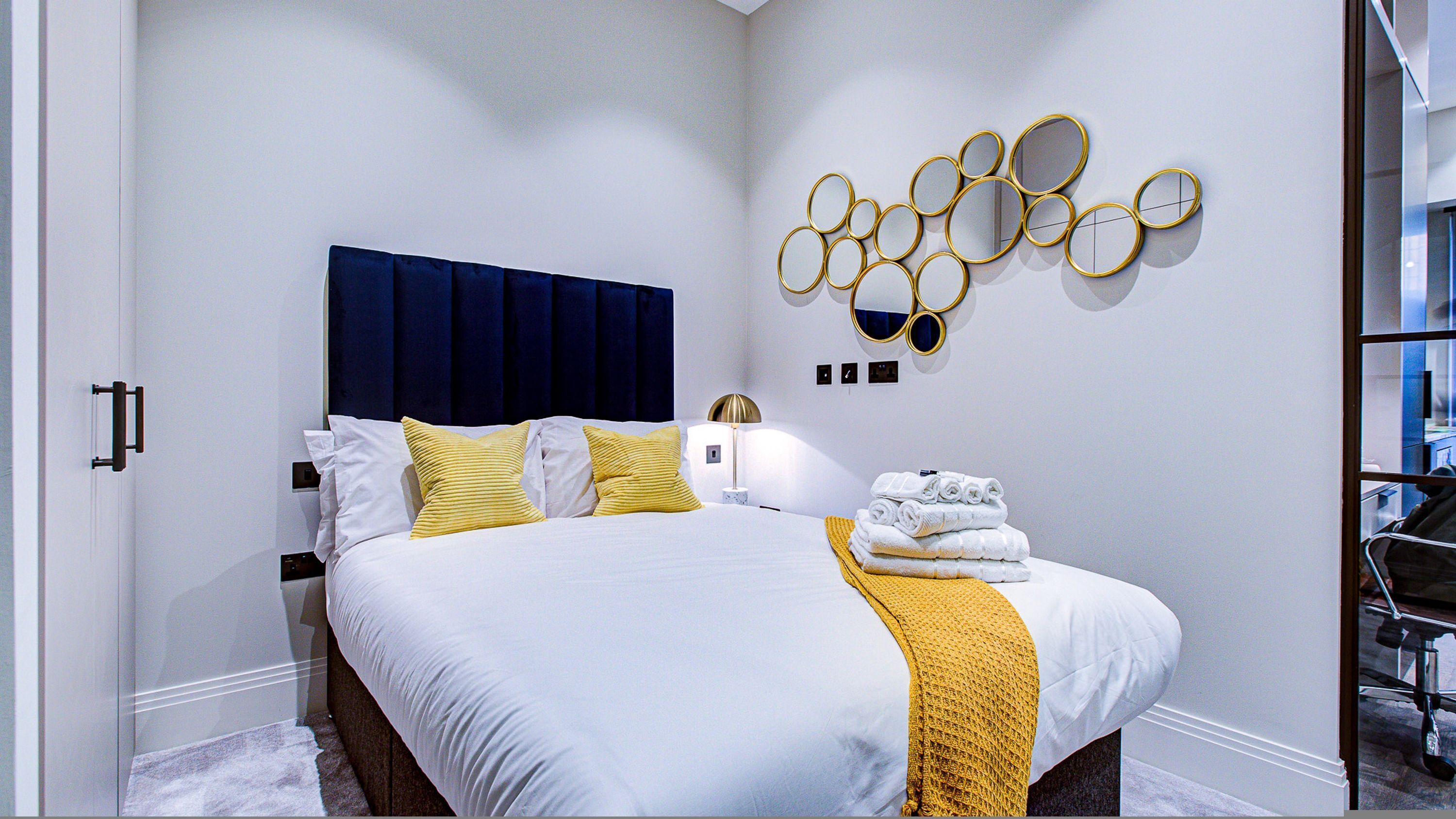 A hotel room with a bed, a headboard, a wall-mounted mirror, and a stack of towels.