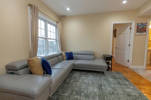 Ein Wohnzimmer mit einem grauen Ecksofa, einem kleinen schwarzen Beistelltisch und einem Gemälde von Boston an der Wand.