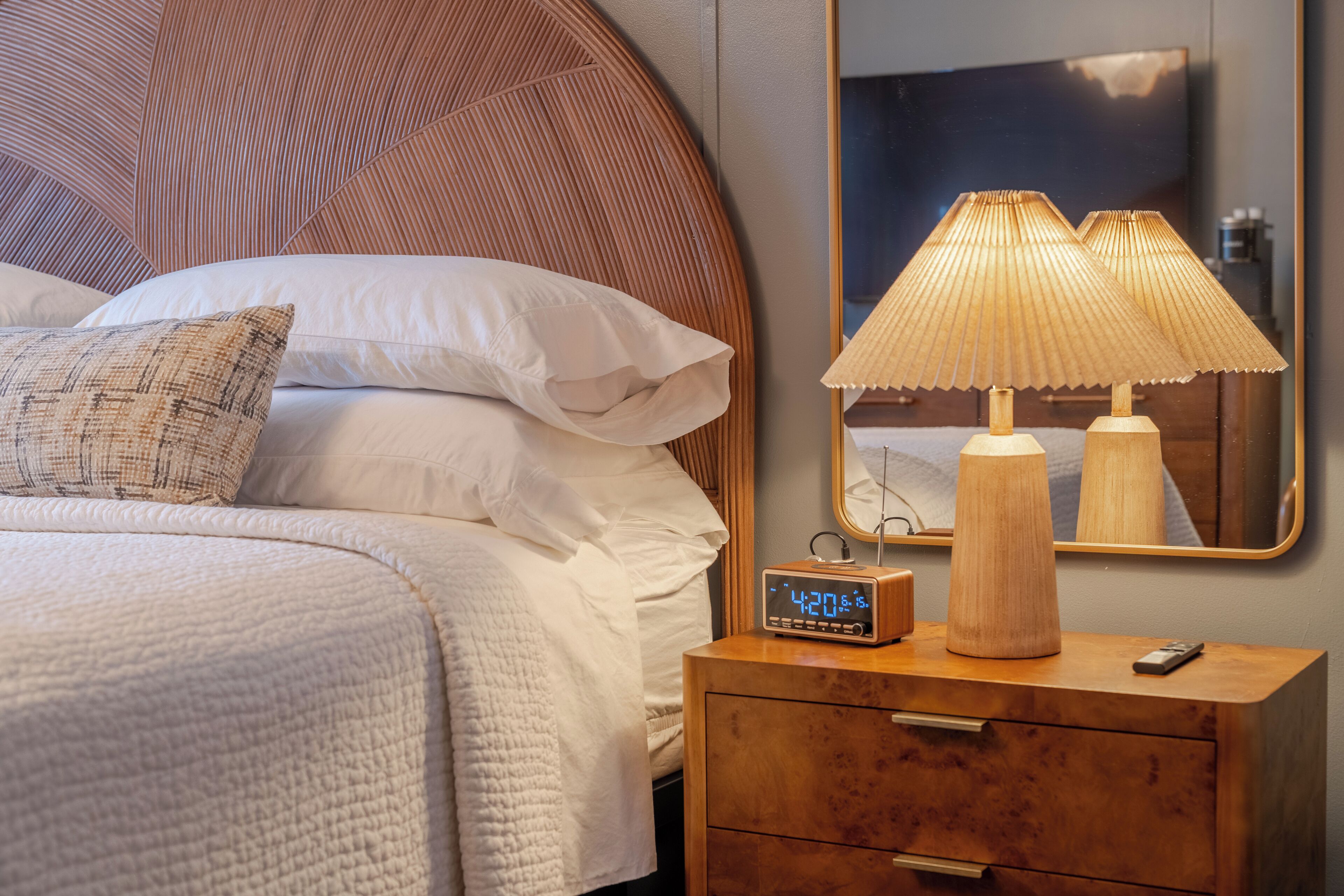 Un lit bien fait, avec du linge de lit blanc et un seul coussin décoratif. Une table de chevet en bois, sur laquelle se trouvent une lampe et une horloge numérique, est placée à côté du lit.