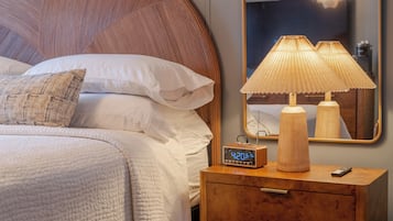 Un lit bien fait, avec du linge de lit blanc et un seul coussin décoratif. Une table de chevet en bois, sur laquelle se trouvent une lampe et une horloge numérique, est placée à côté du lit.