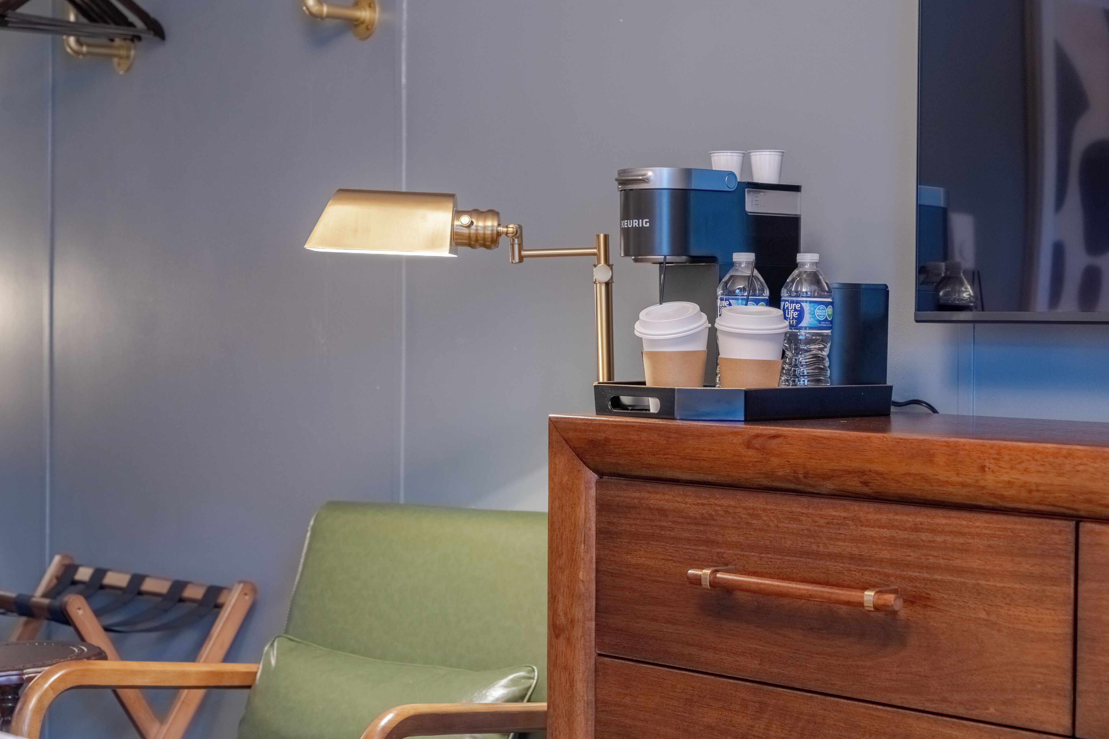 A hotel room with a wooden dresser, a green chair, a desk with a coffee machine, and a lamp.