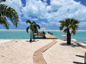 Ein hölzerner Weg führt zu einem Strandbereich mit einer Holzterrasse, Palmen und freiem Blick auf den Ozean.