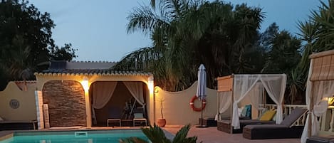 Una piscina con un macetero de palmera en primer plano, rodeada de sillas de descanso y una zona de estar cubierta.