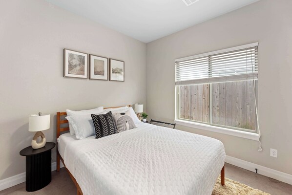 A bedroom with a bed, bedside tables, a window with blinds, and three framed pictures on the wall.