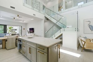 Eine moderne Küche mit einer zentralen Kochinsel, einem Essbereich mit Holztisch und einer Treppe mit Glastrennwand.