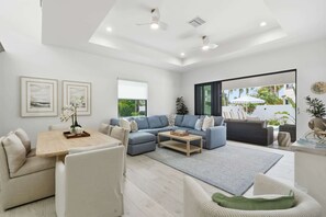 A modern living room with a sectional sofa, a dining table, and a large window. - Brand-New Luxury Pool Home Steps from the Gulf (Naples)