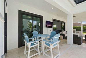 An outdoor patio with a round table and chairs, a television mounted on the wall, and a view of the outdoors. - Brand-New Luxury Pool Home Steps from the Gulf (Naples)