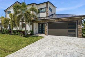 A modern two-story house with a garage, a well-maintained lawn, and a variety of tropical plants. - Brand-New Luxury Pool Home Steps from the Gulf (Naples)