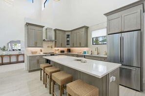 A modern kitchen with a large island, stainless steel appliances, and a view to the outdoors. - Brand-New Luxury Pool Home Steps from the Gulf (Naples)