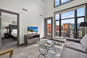 A modern hotel room with a bed, a flat-screen TV, a coffee table, and large windows offering a view of buildings and a beach. - 2 Bd Loft 603 at Playa. Pool and Hot Tub (Kelowna)