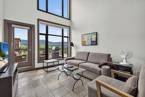 A spacious living room with a large sliding glass door leading to a balcony, a comfortable sofa, a glass coffee table, and a television. - 2 Bd Loft 603 at Playa. Pool and Hot Tub (Kelowna)