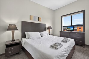 A hotel room with a bed, nightstands, a TV, and a view of buildings through a window. - 2 Bd Loft 603 at Playa. Pool and Hot Tub (Kelowna)