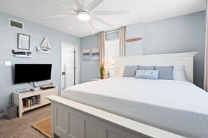 A bedroom with a large bed, a ceiling fan, a TV, a bookshelf, and a door. - Santa Rosa Dunes 321 (Pensacola Beach)
