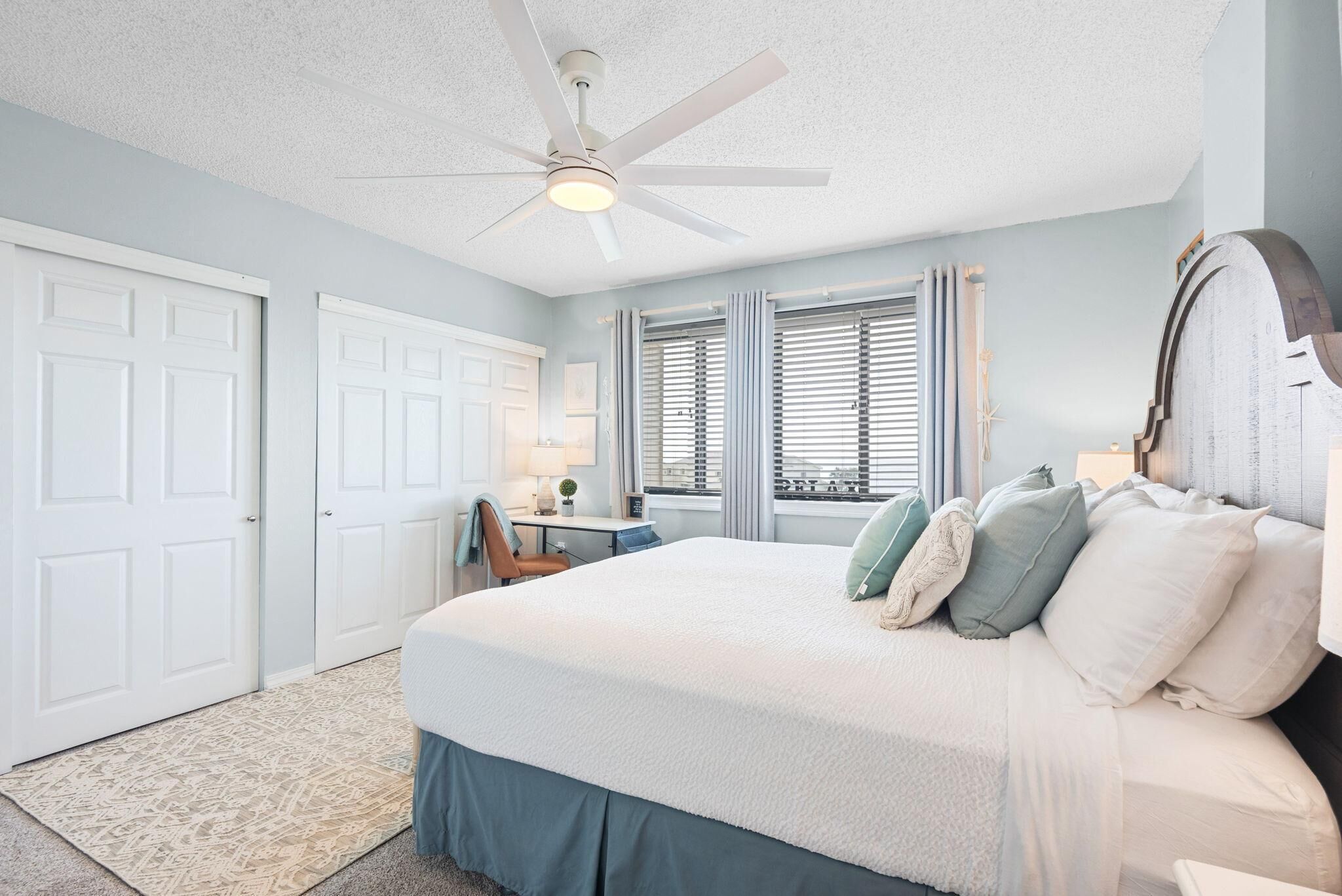 A bedroom with a large bed, a ceiling fan, a desk with a chair, and a window with blinds.