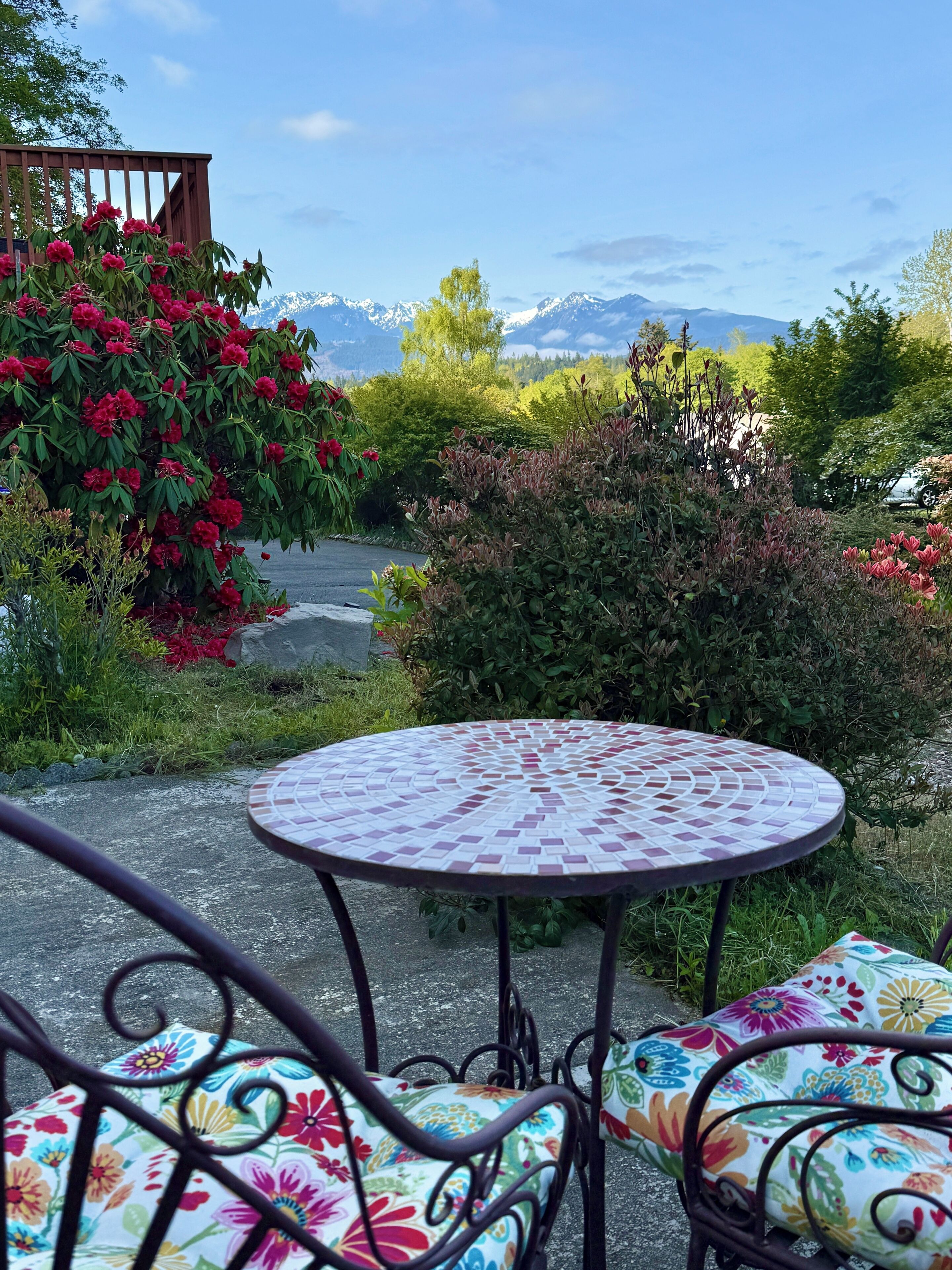 Une table ronde d’extérieur, ornée d’un plateau en mosaïque, entourée de chaises en métal aux motifs floraux, le tout se fondant dans un décor de verdure luxuriante et de montagnes lointaines.