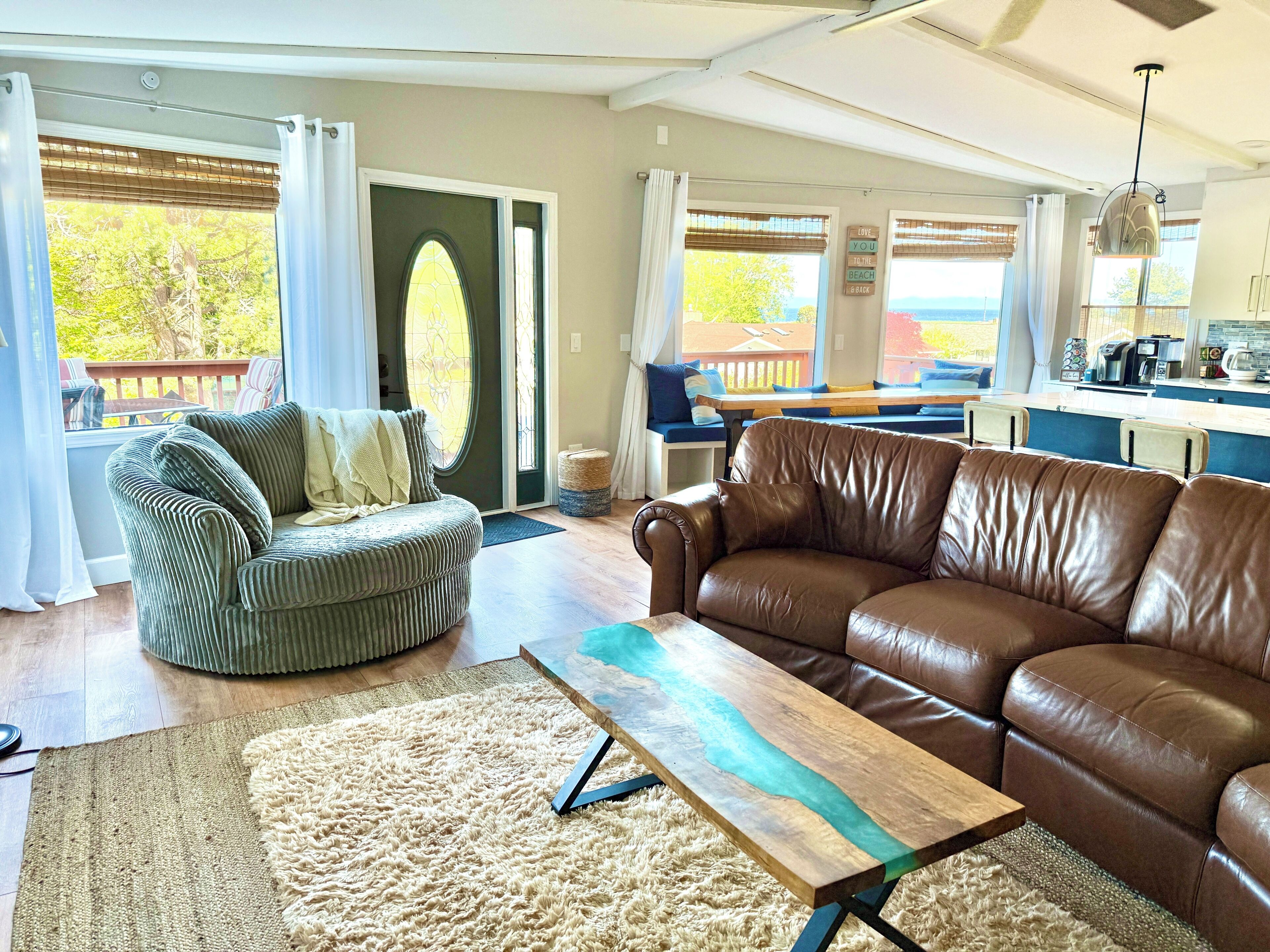Ein Wohnzimmer mit einem braunen Ledersofa, einem grünen Polsterstuhl, einem Holztisch und Blick ins Freie durch große Fenster.