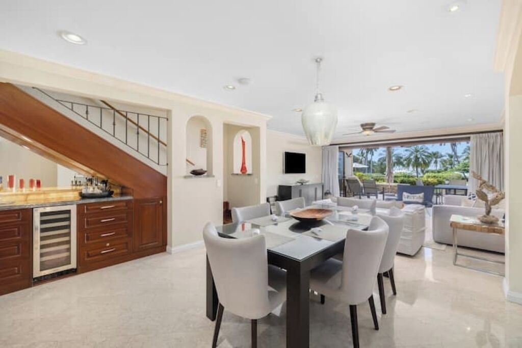 A modern dining area with a large table and chairs, a kitchen with a wooden staircase, and a view of palm trees through large windows.