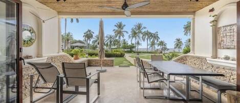 Une terrasse extérieure avec une table et des chaises, un ventilateur de plafond, et une vue sur des palmiers et un ciel dégagé.
