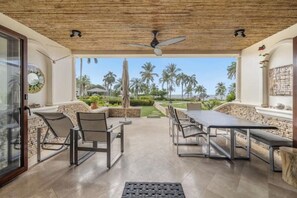 Eine Terrasse im Freien mit Esstisch und Stühlen, einem Deckenventilator sowie Blick auf Palmen und einen klaren Himmel.