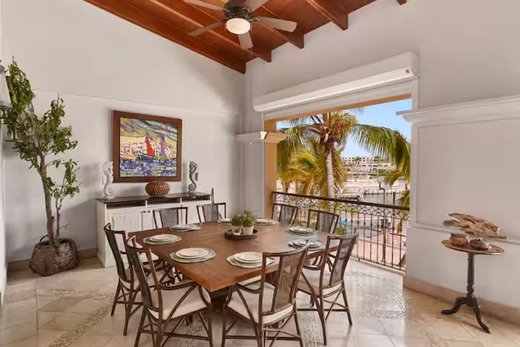 A dining area with a wooden table set for six, surrounded by chairs, with a view of a pool and palm trees outside.