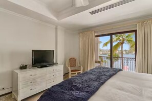 Hotelkamer met een bed, een televisie op een dressoir, een stoel en een balkon met uitzicht op palmbomen en een wateroppervlak.