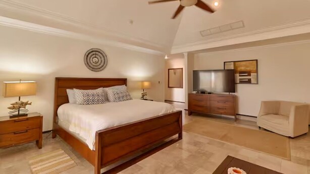 Une chambre à coucher comprenant un lit, des lampes de chevet, un ventilateur de plafond, une commode, un téléviseur et une chaise.