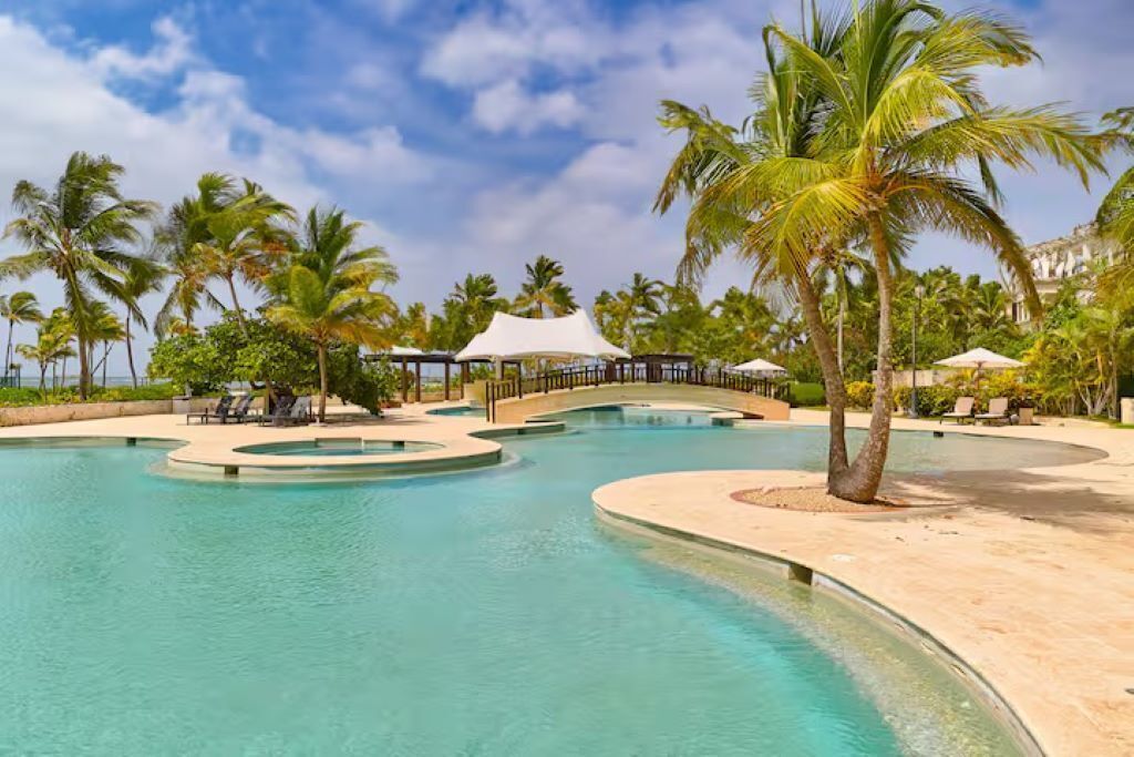 Un espace piscine aménagé dans un complexe hôtelier, avec des palmiers, des chaises longues et un coin salon abrité.