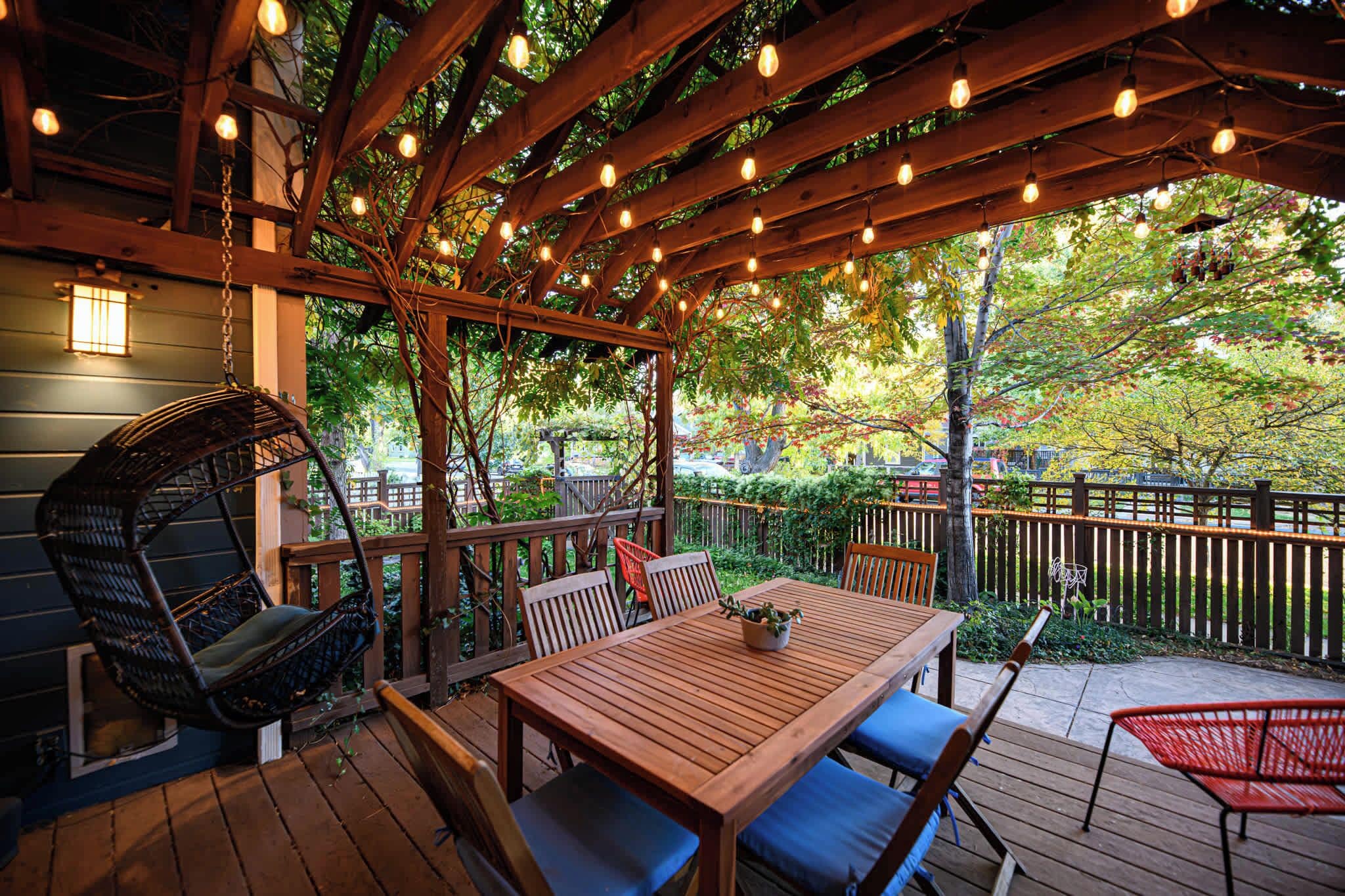 Une terrasse en bois avec une table et des chaises, un fauteuil suspendu et une pergola illuminée de guirlandes lumineuses.