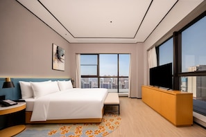 Habitación de hotel con una cama grande, un televisor sobre un mueble de madera y vistas a la ciudad a través de amplias ventanas.
