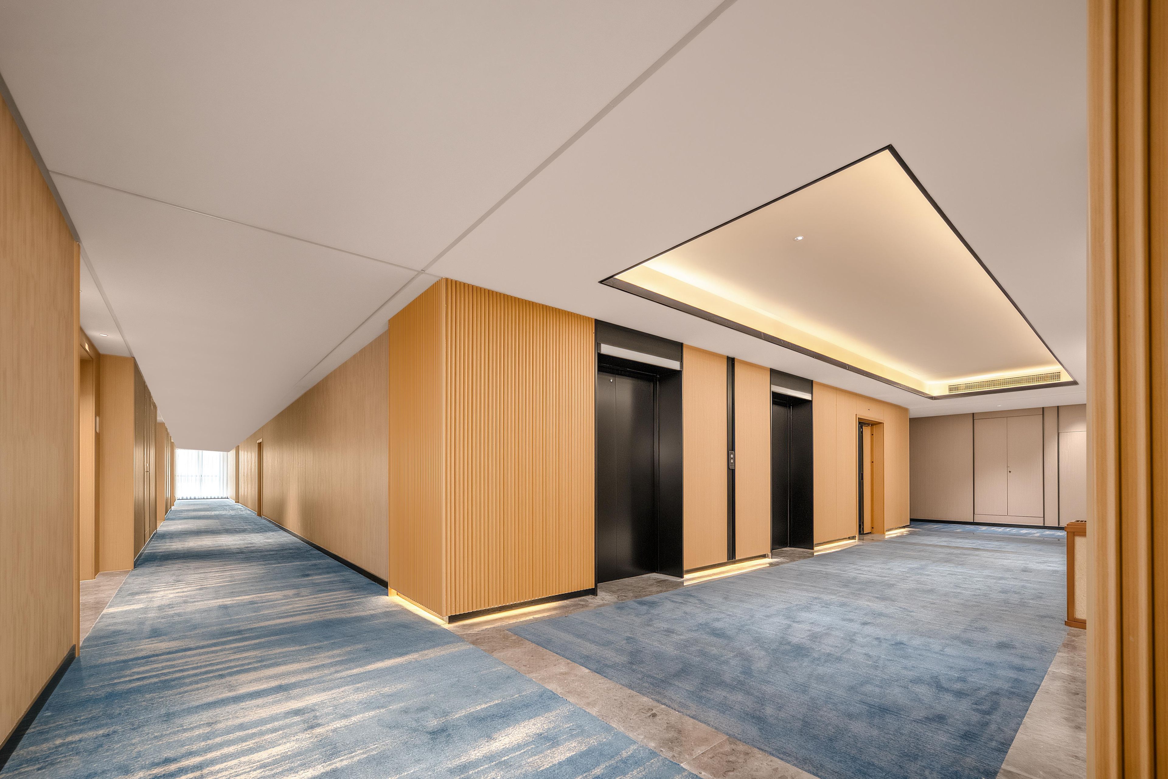 A modern hotel corridor with a blue carpet, wooden panel walls, and a ceiling with recessed lighting.
