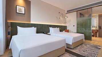 A hotel room with two beds, a wall-mounted artwork, a bedside table with a phone, and a glass partition leading to a bathroom.