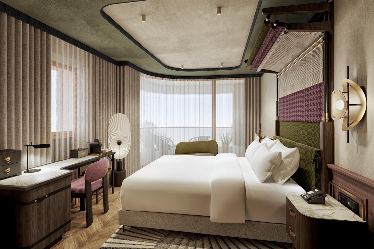 A modern hotel room with a large bed, a desk, and a chair. There is a window with curtains, a ceiling fan, and a wall-mounted lamp.