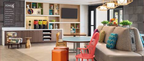 A modern hotel room with a sofa, chairs, a dining table, a kitchen area, and a TV.