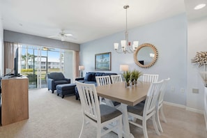 A modern living room with a dining area, a ceiling fan, a chandelier, and a large window with curtains. - 414 - Green Links First Floor -Sleeps 6 (Naples)