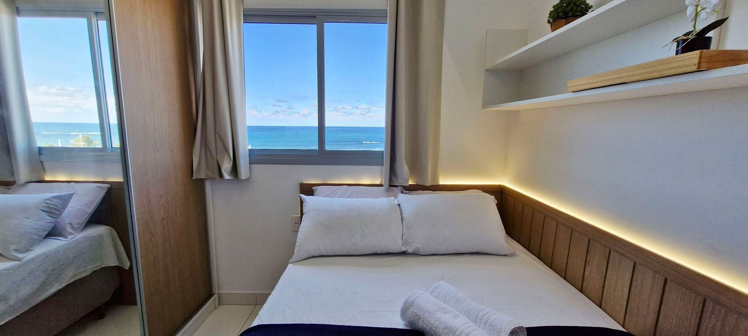 Habitación de hotel con cama, una cama más pequeña, ventana con vista al mar y estante con una planta.