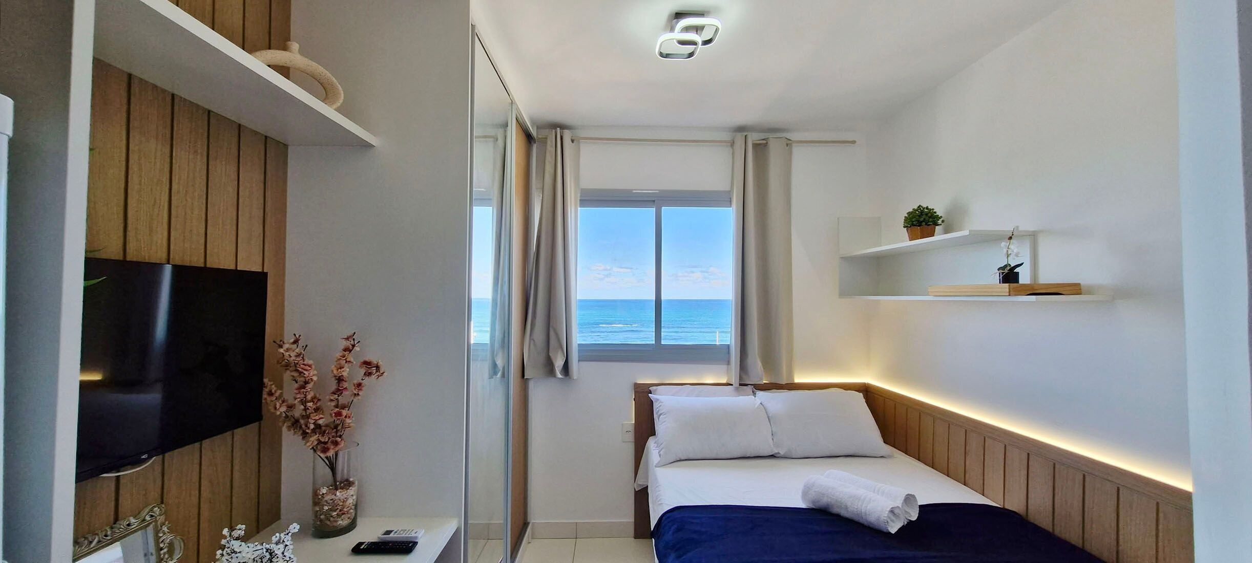 A hotel room with a bed, a TV, a small table with a vase of flowers, and a window with a view of the sea.