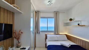 Quarto de hotel com cama, televisão, uma mesinha com um vaso de flores e janela com vista para o mar.