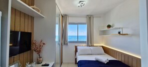 A hotel room with a bed, a TV, a small table with a vase of flowers, and a window with a view of the sea. - COPIA0108 - Edifício Essencial Piata (Salvador)