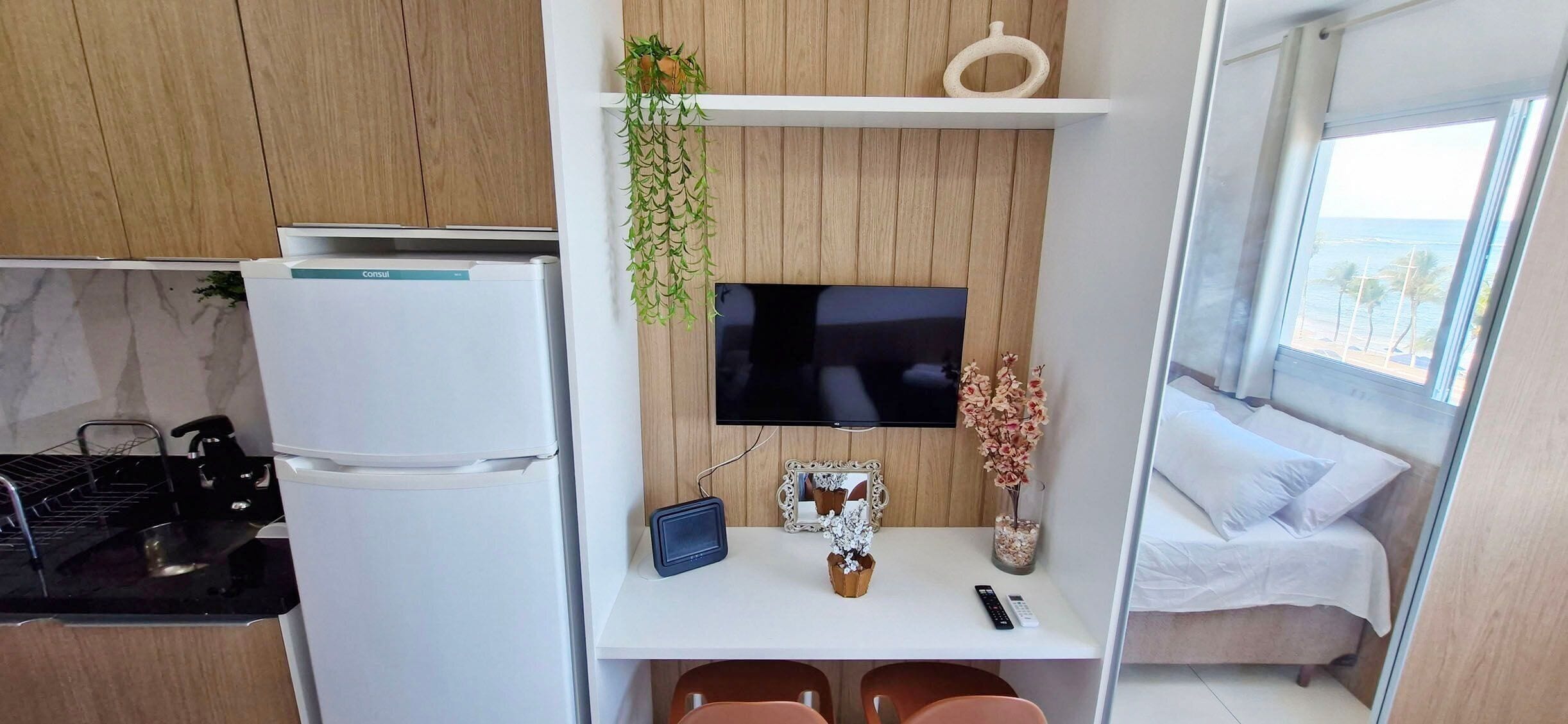 Una kitchenette compacta con refrigerador, estufa y lavamanos. Un televisor de pantalla plana está montado en una pared de madera sobre una pequeña mesa con una tableta, un jarrón y un control remoto. Se ve una cama con ropa blanca a través de una ventana.